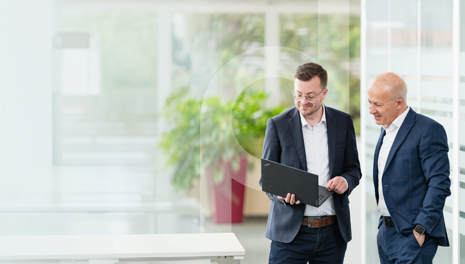 Two businessmen are standing in a modern office building, discussing something on a laptop with digital overlays.
