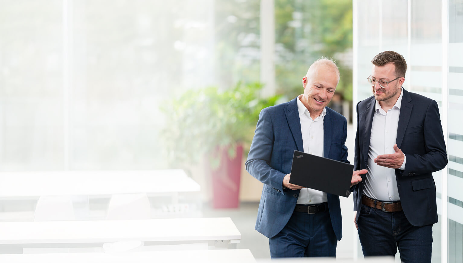 Zwei Männer in Business-Kleidung stehen in einem hellen Büro und besprechen etwas auf einem gemeinsam gehaltenen Laptop.