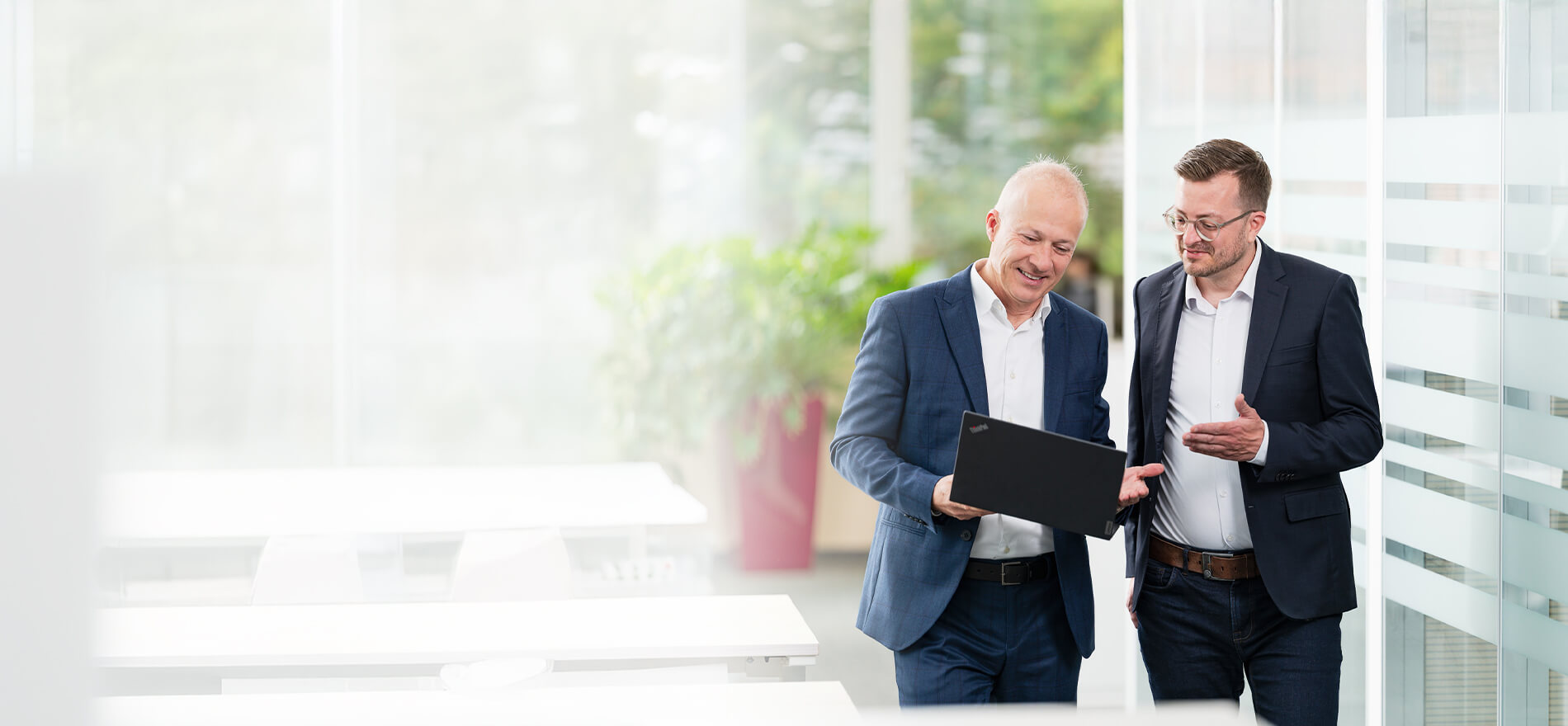 Two men in business attire are standing in a bright office discussing something on a laptop they are holding together.