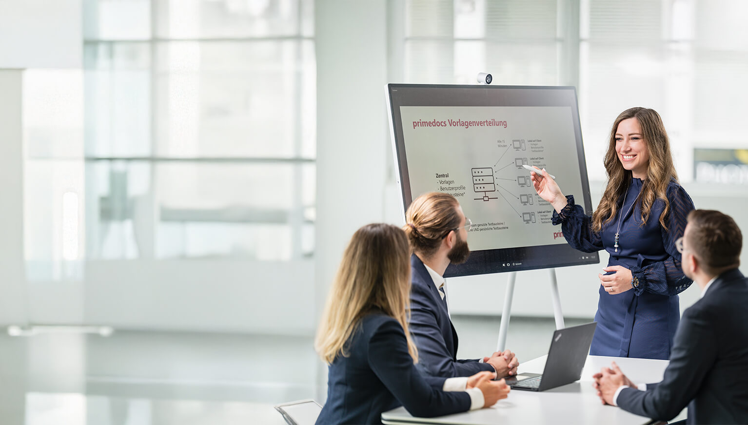A businesswoman gives a presentation to four colleagues in the meeting room and points to a digital whiteboard.