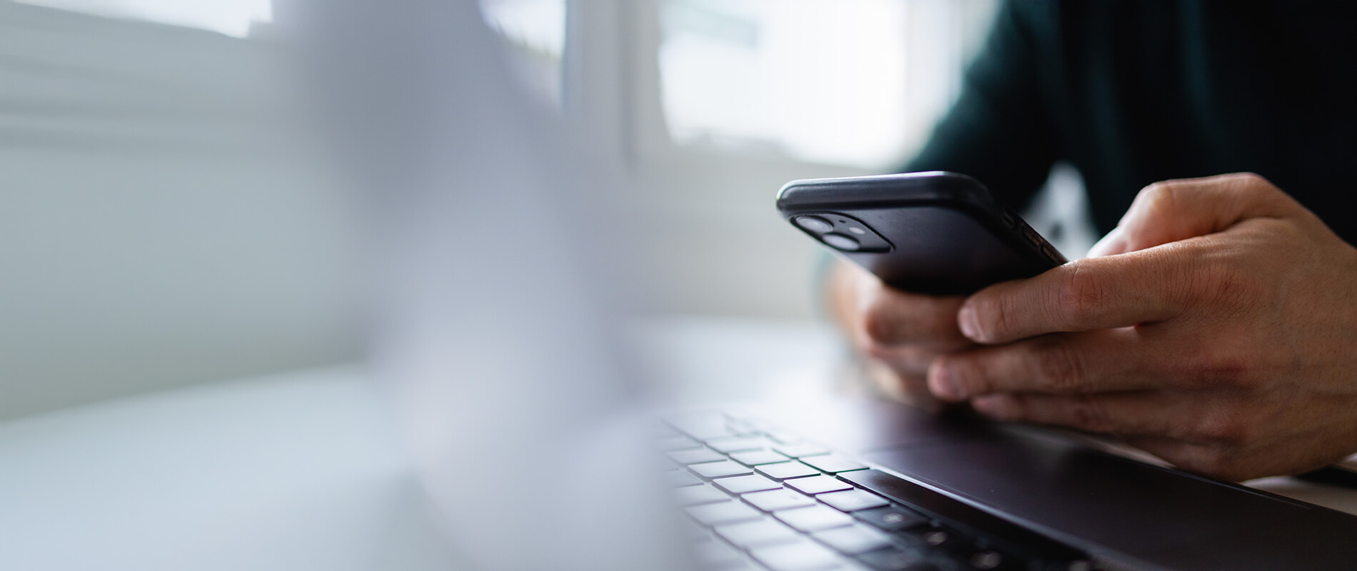 Close-up of hands holding and operating a smartphone above a laptop keyboard.