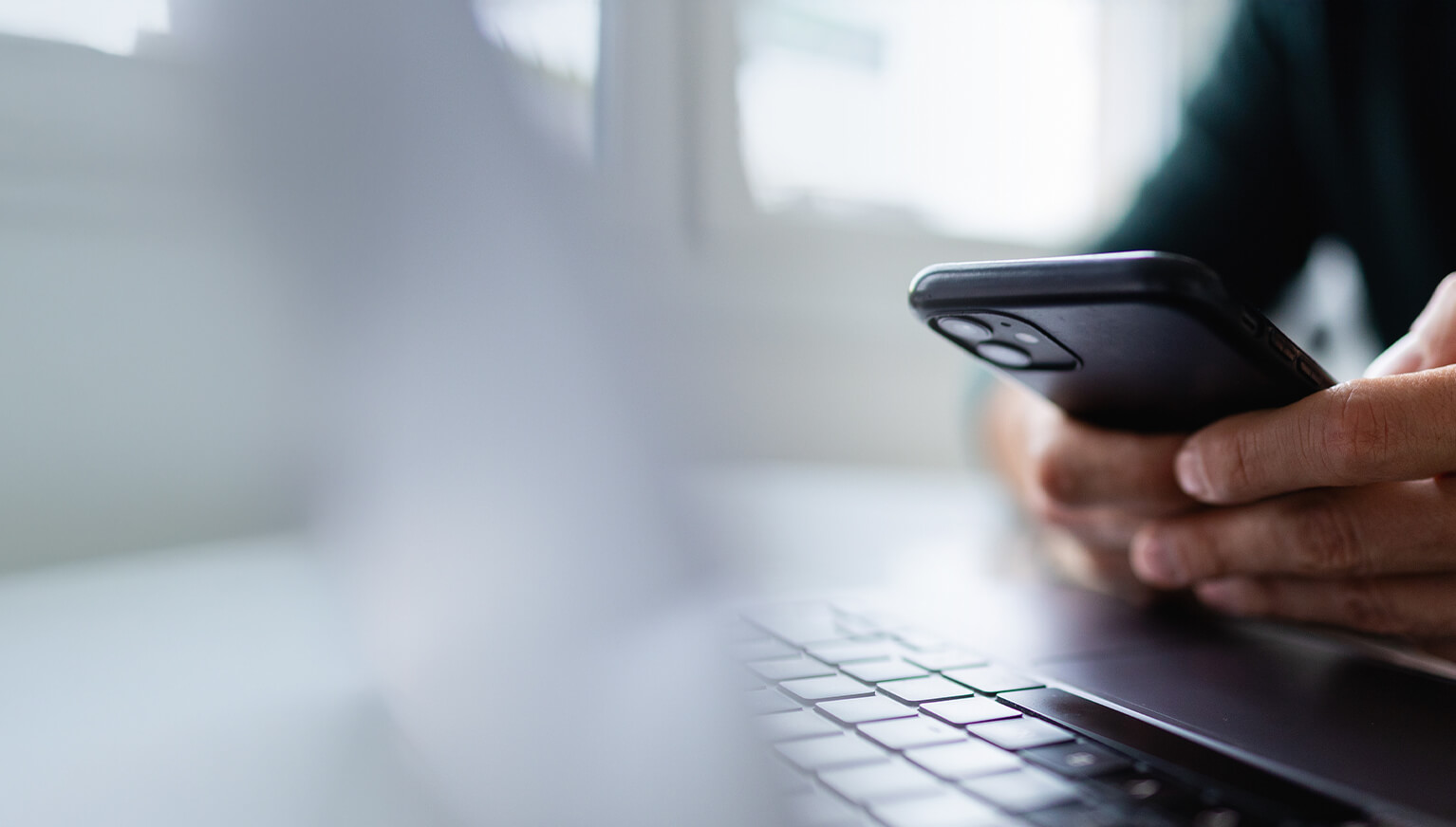 Close-up of hands holding and operating a smartphone above a laptop keyboard.