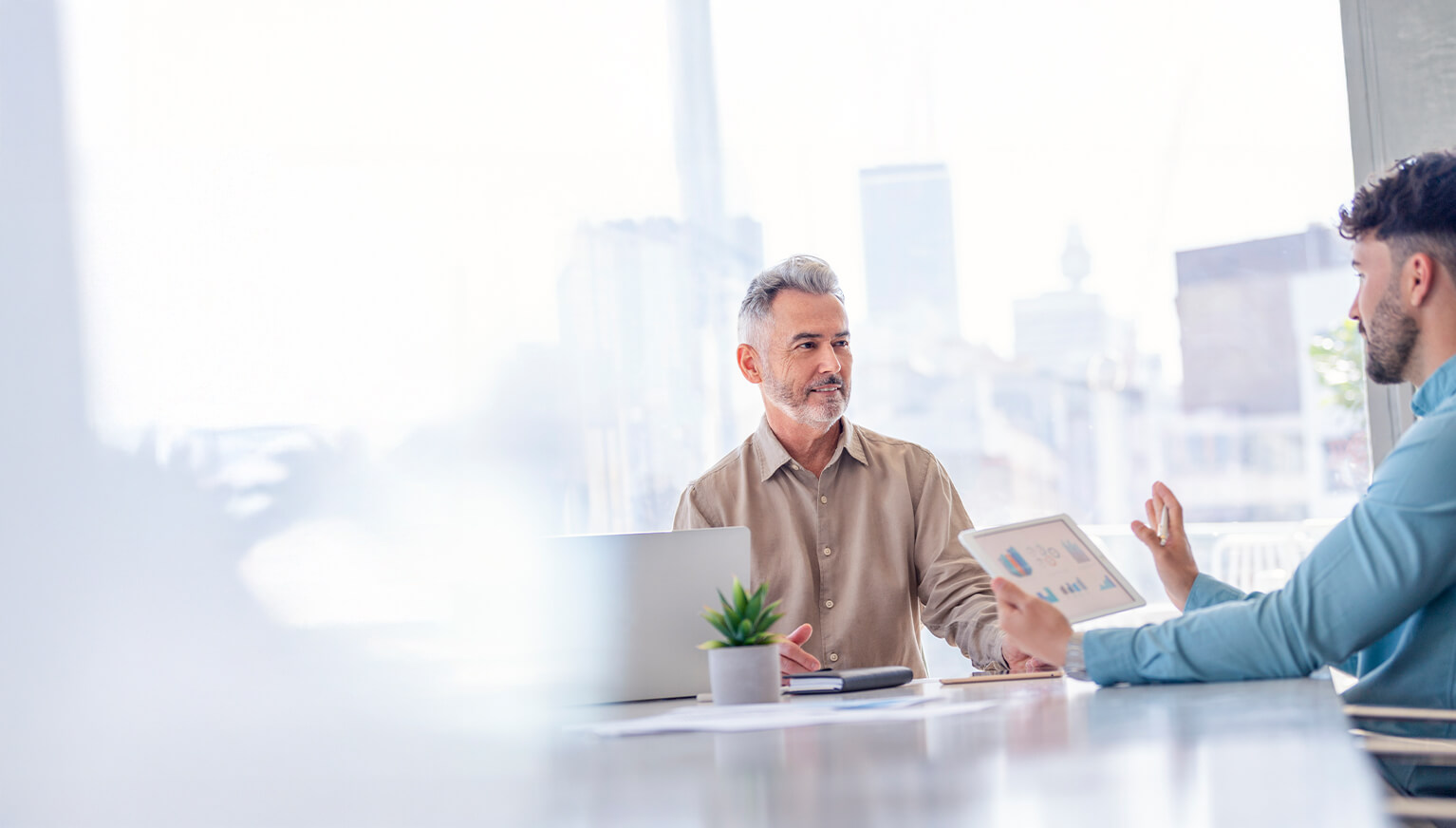 Zwei Männer in einem modernen Büro mit großer Fensterfront besprechen Geschäftszahlen auf einem Tablet.