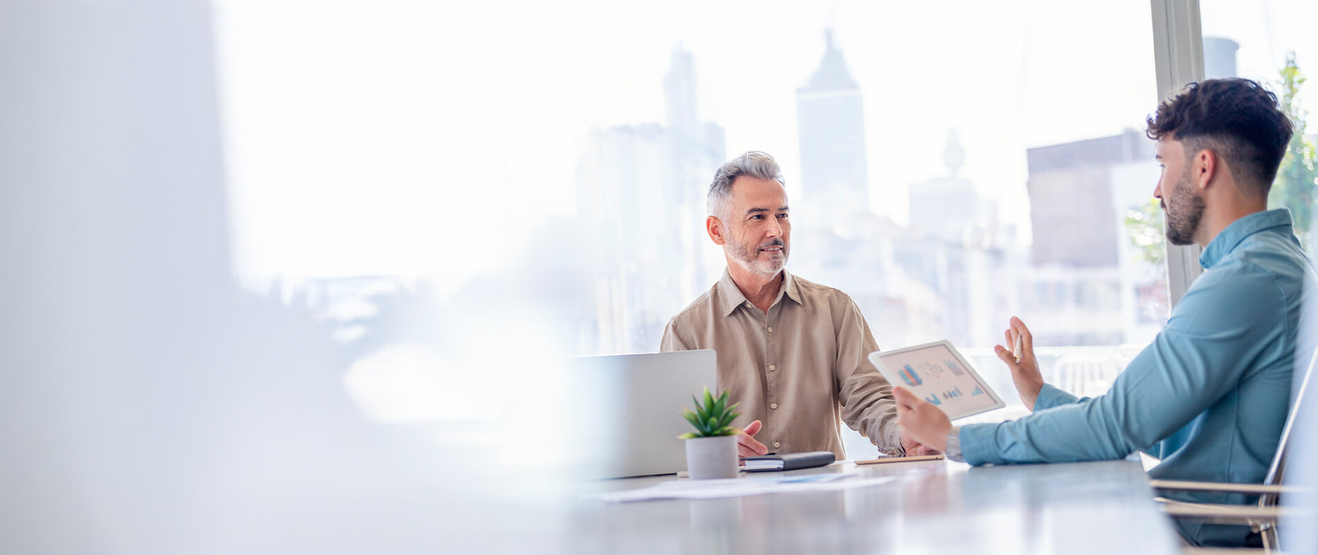 Two men in a modern office with large windows discuss business figures on a tablet.
