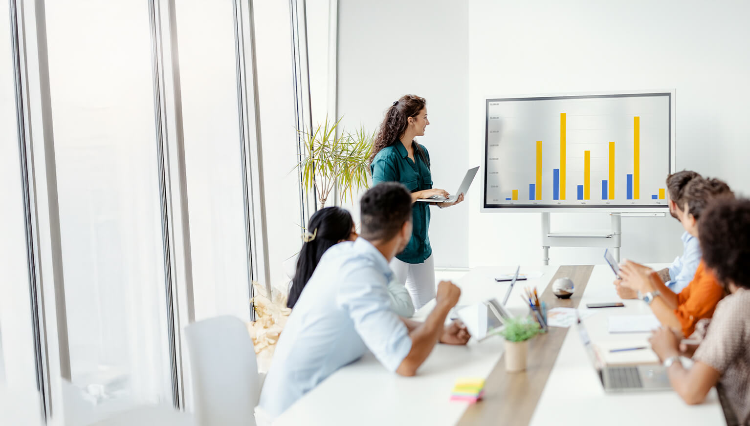 Eine junge Frau präsentiert Geschäftszahlen mit einem Laptop auf einem großen Bildschirm vor einem Team in einem Meetingraum.