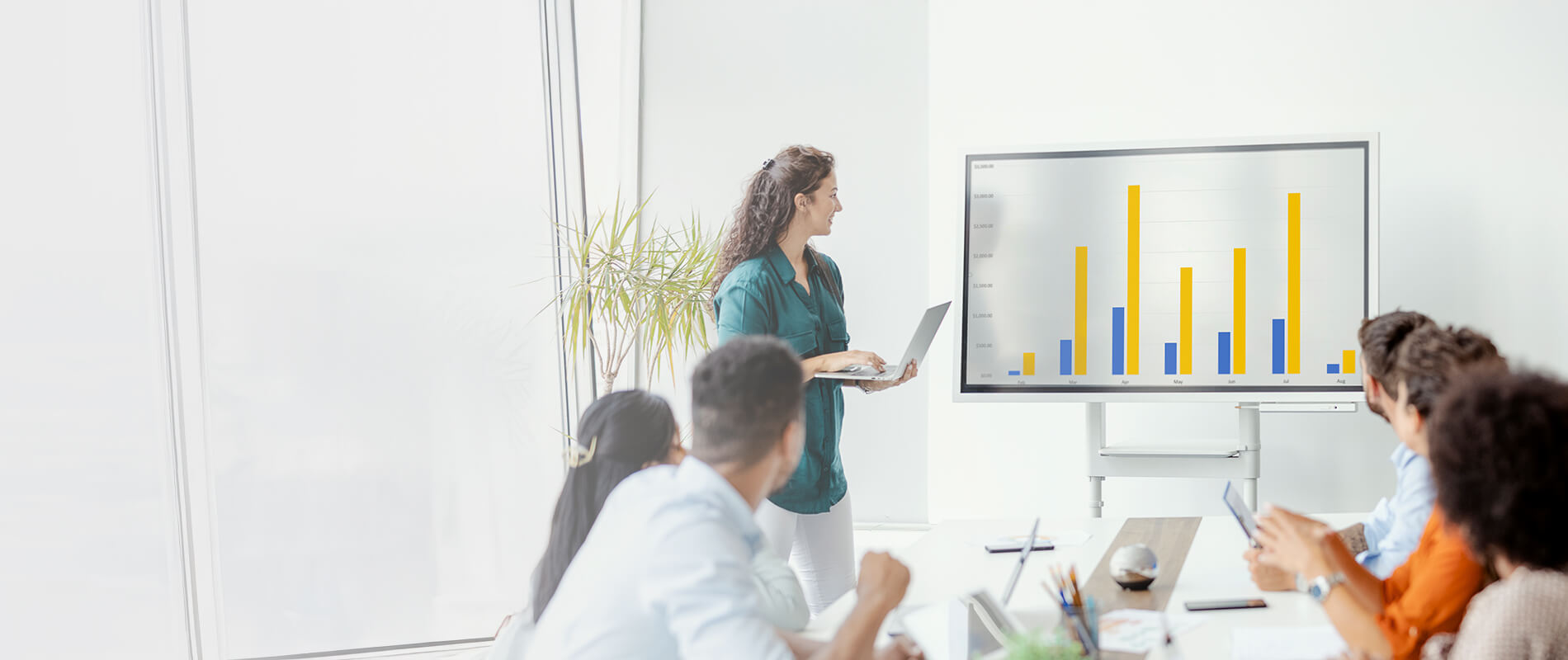Eine junge Frau präsentiert Geschäftszahlen mit einem Laptop auf einem großen Bildschirm vor einem Team in einem Meetingraum.