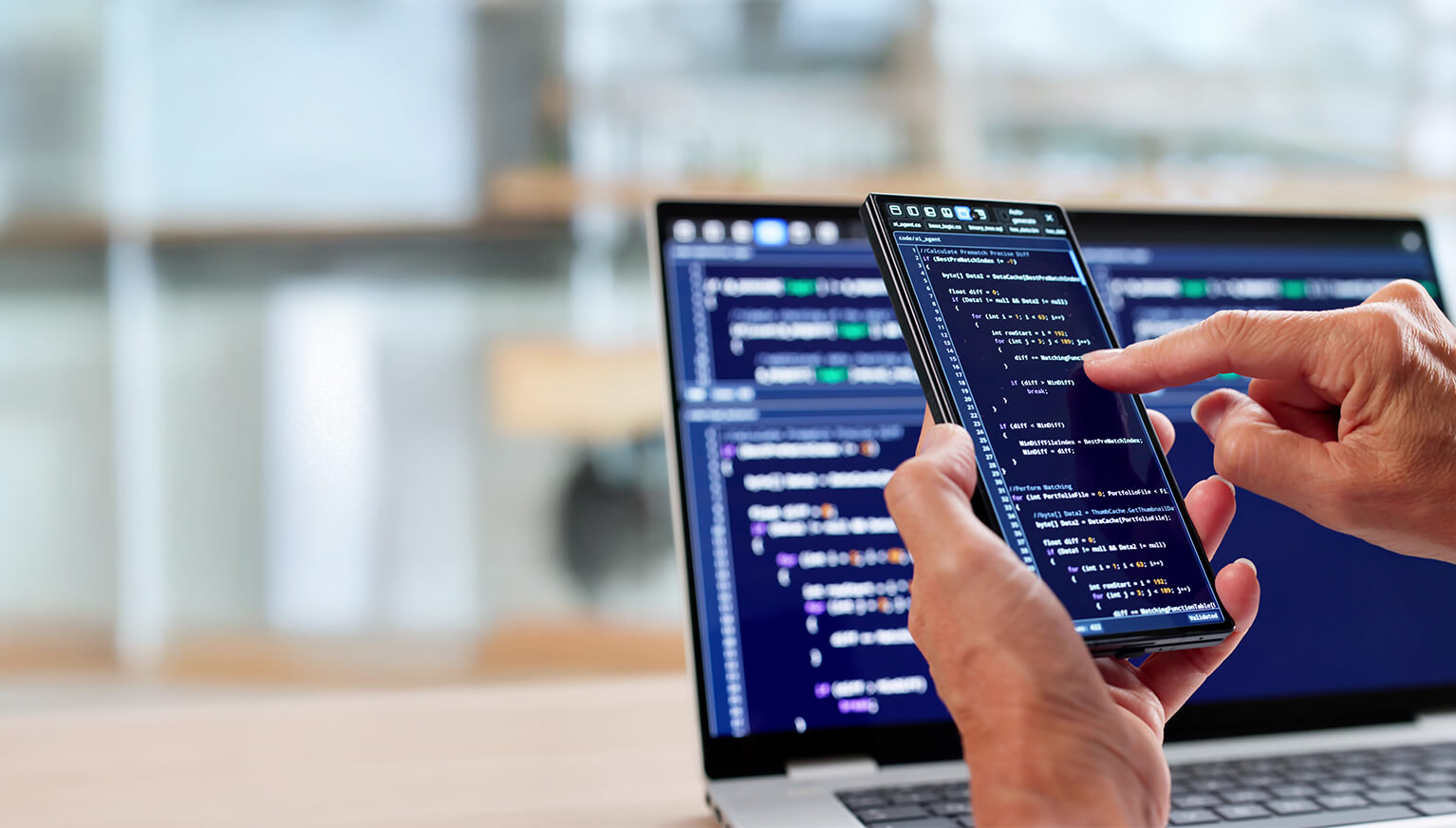 Hands typing code on a smartphone, whose screen is placed in front of a laptop with more code in the office.