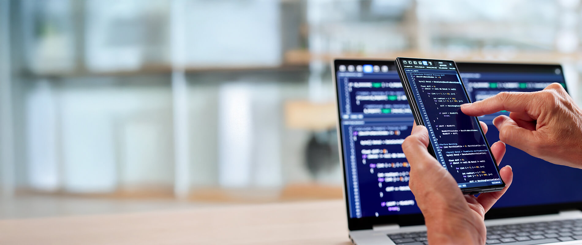 Hands typing code on a smartphone, whose screen is placed in front of a laptop with more code in the office.