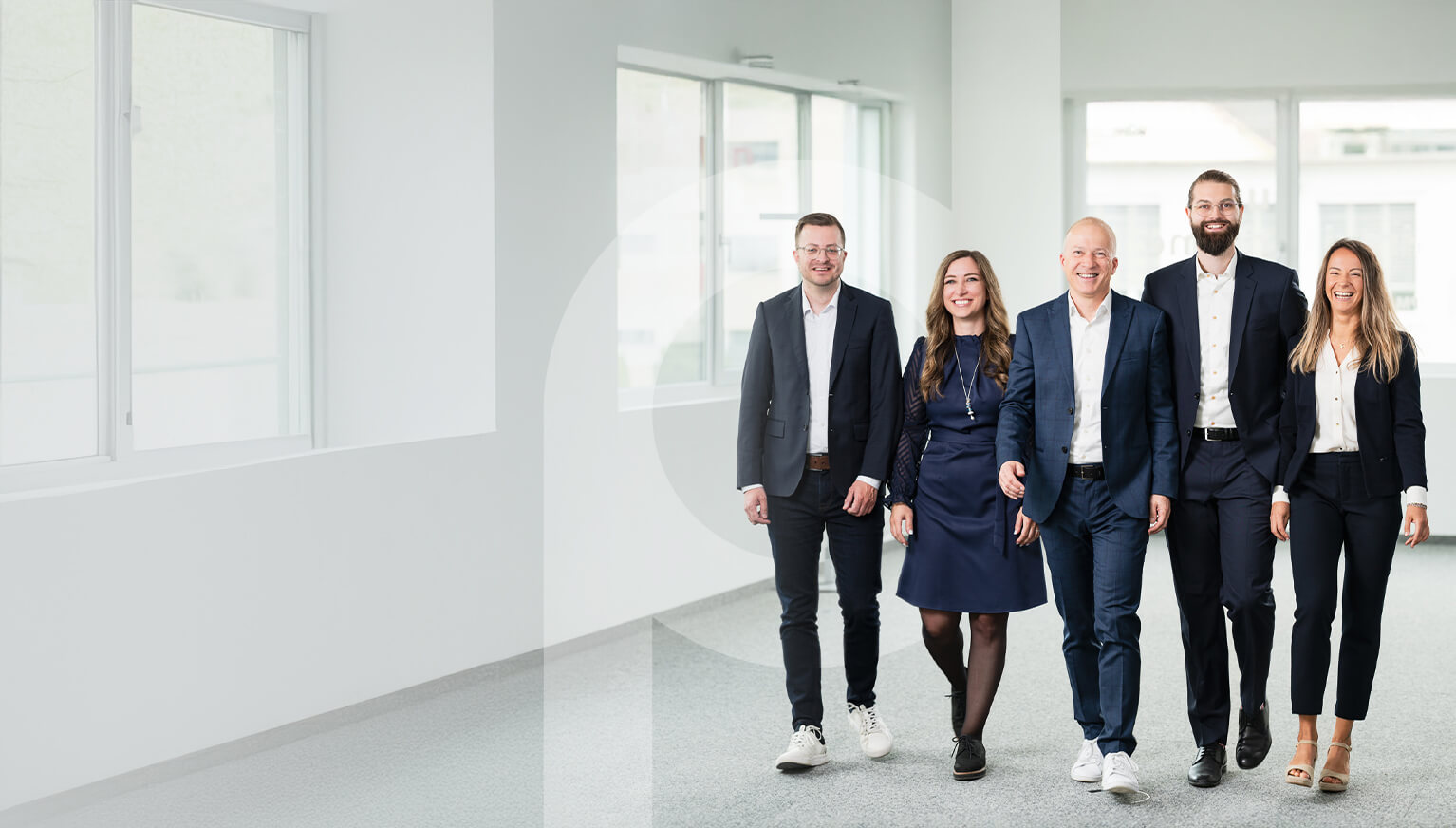Five smiling business professionals from the PrimeSoft Group, dressed in business attire, walk together through a bright, modern office corridor.