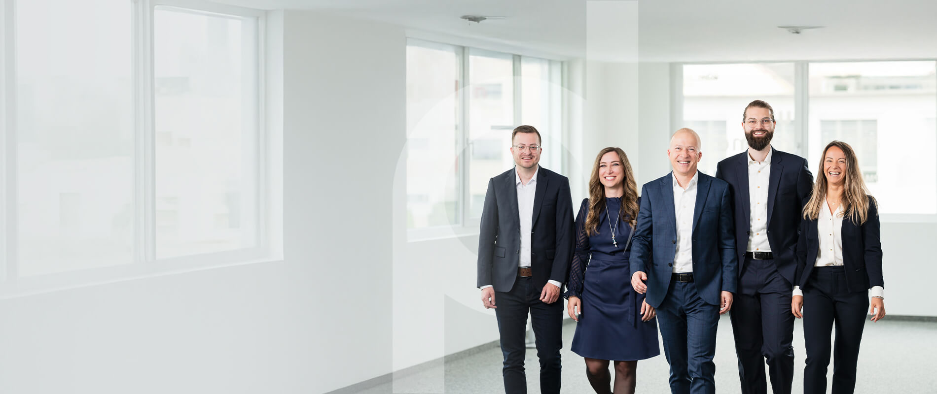 Five smiling business professionals from the PrimeSoft Group, dressed in business attire, walk together through a bright, modern office corridor.