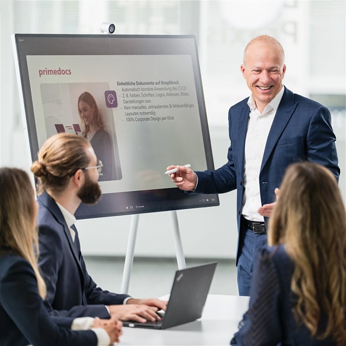 A smiling businessman gives a presentation to three colleagues using a digital whiteboard.
