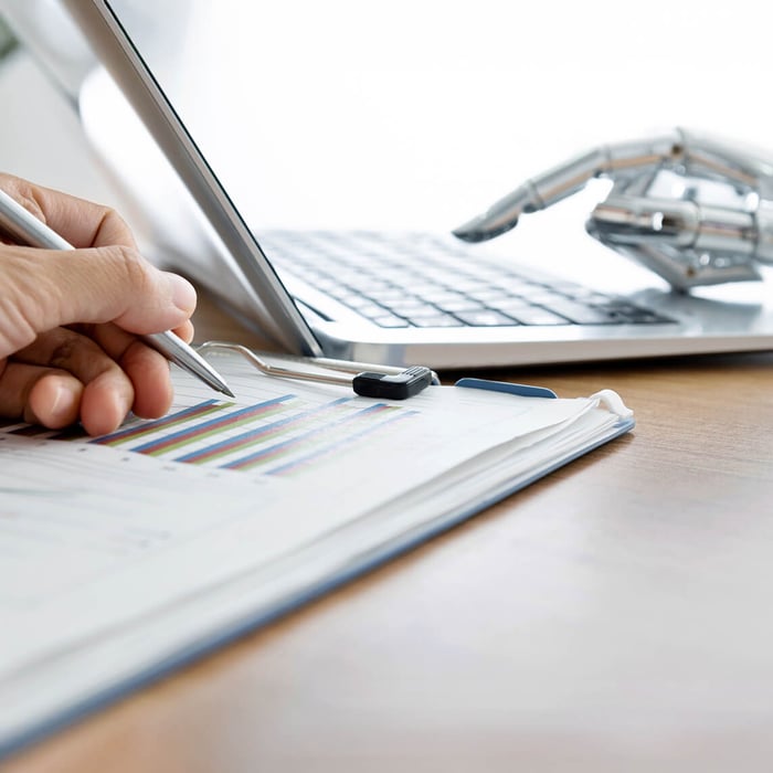 A human hand writes on a diagram with a pen. In the background, a robotic hand types on a laptop.