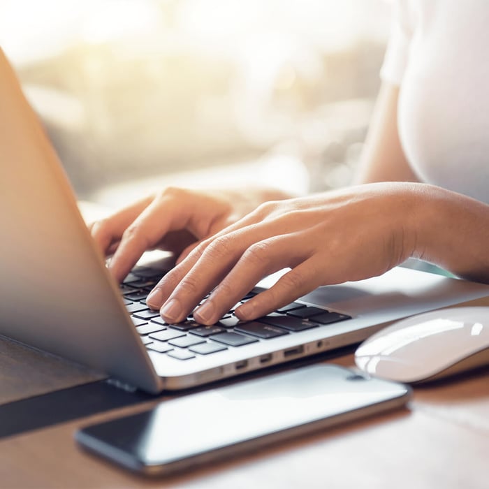 Close-up of hands typing on a laptop keyboard, with a smartphone and mouse in the foreground. 