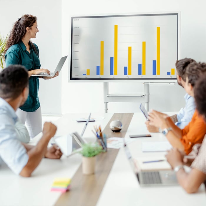 A young woman presents business figures using a laptop on a large screen in front of a team in a meeting room.