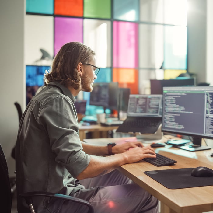 Entwickler arbeitet konzentriert an zwei Monitoren mit Code in einem modernen Büro mit bunter Glaswand.