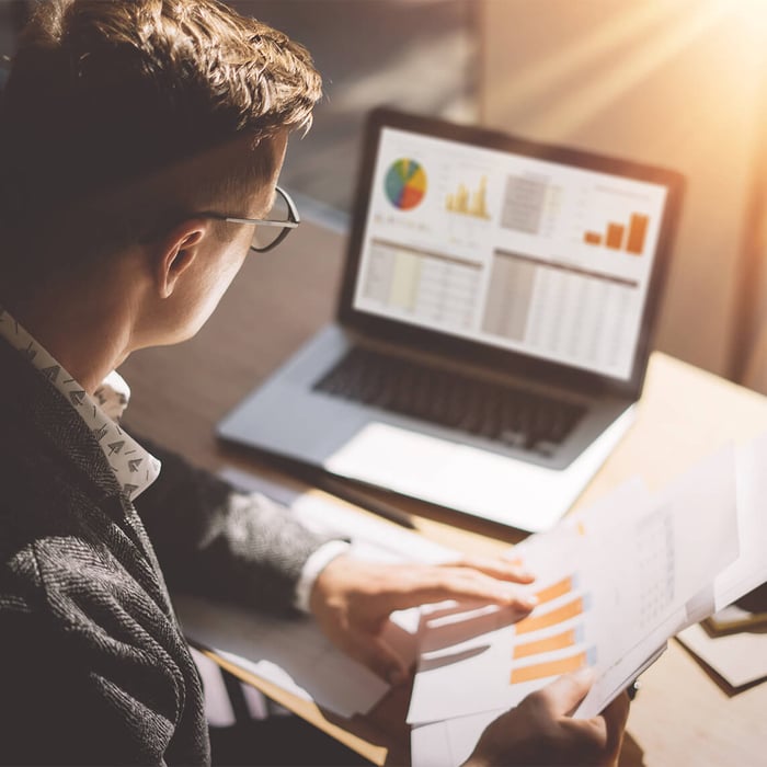 Man reading documents at his desk. In the background, his laptop displays business charts and graphs (data analysis).