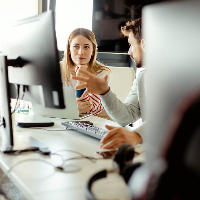 Two colleagues in an office are engaged in an intense discussion at a desk. The woman is listening to the man, who is gesturing.