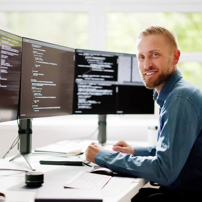 Lachender, bärtiger Entwickler sitzt vor drei Monitoren mit Code. Er trägt ein blaues Hemd.