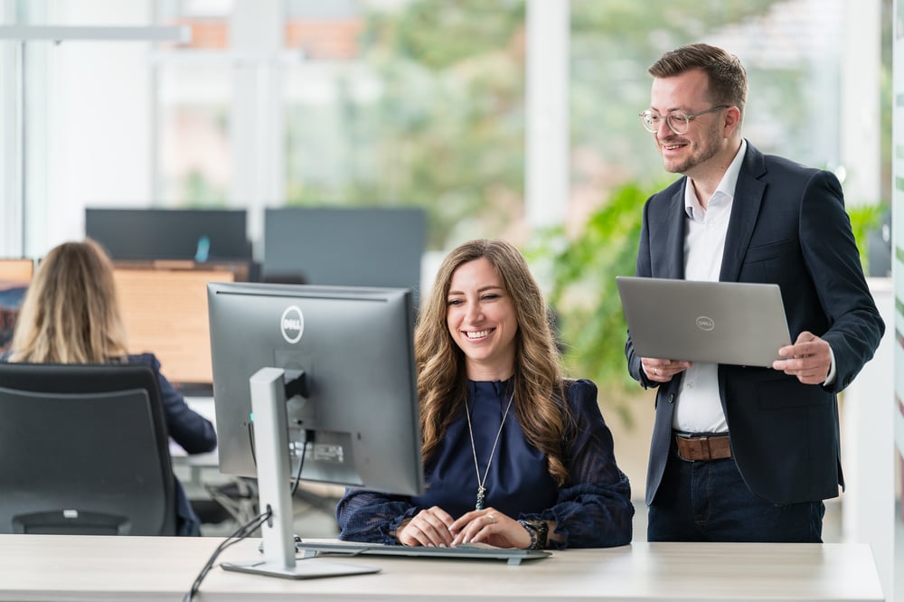 Two smiling colleagues are working on a Dell PC in a modern office. The man is standing next to the woman with a laptop.