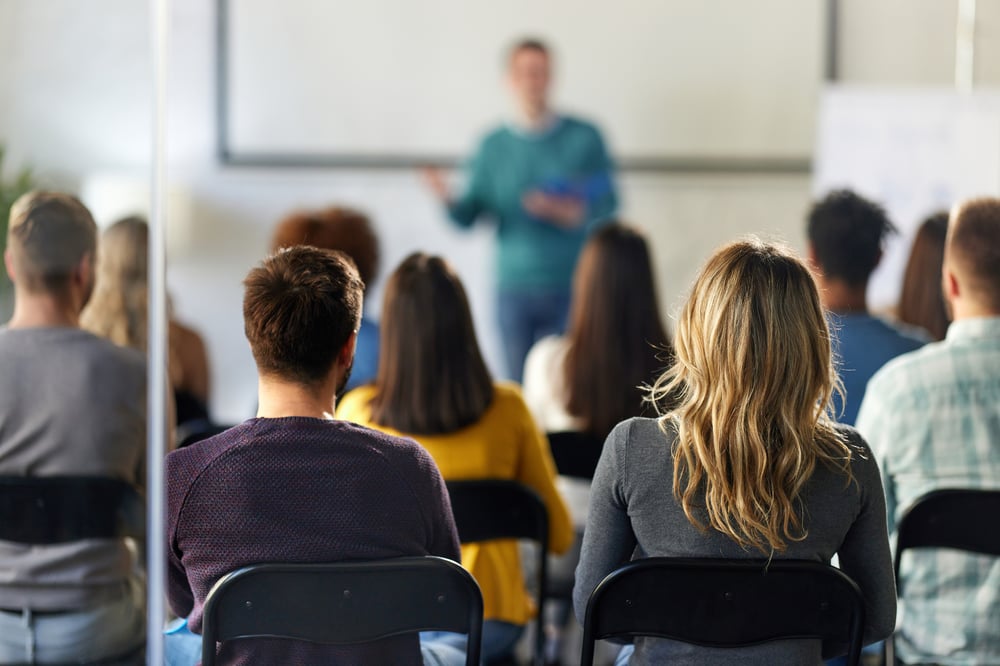 Rear view of a group of listeners in a seminar room listening to a person speaking at the screen.