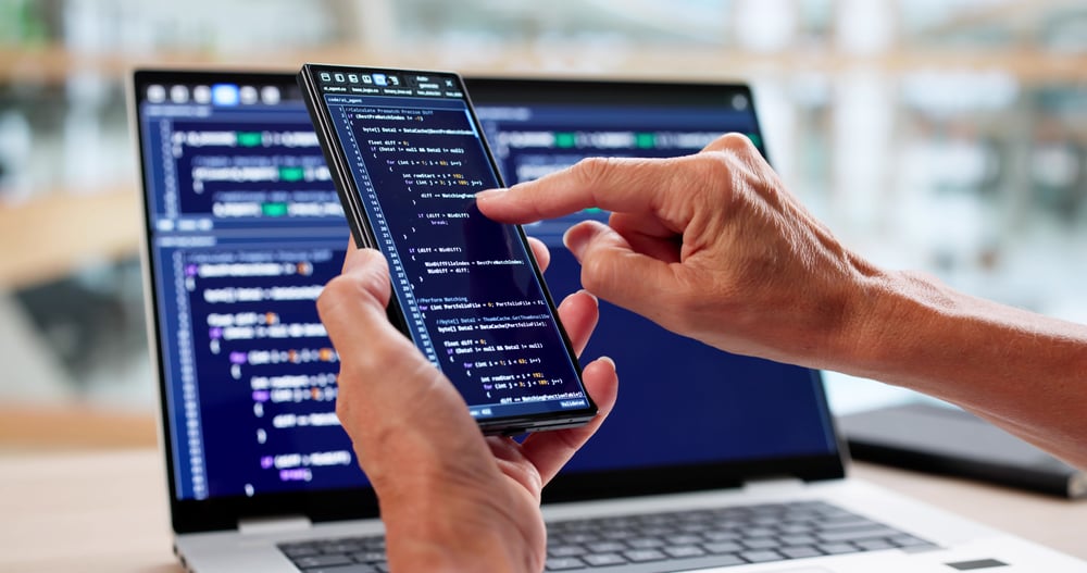 Close-up of hands checking code on a smartphone while code is displayed on a laptop in the background.