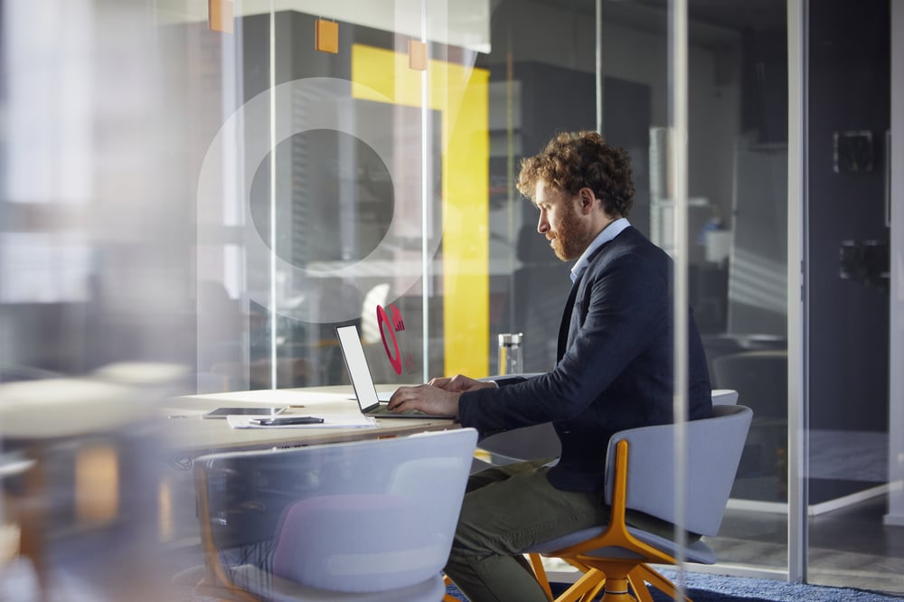 Man with red hair and beard working intently on a laptop in a modern office. A digital element is visible on the screen.