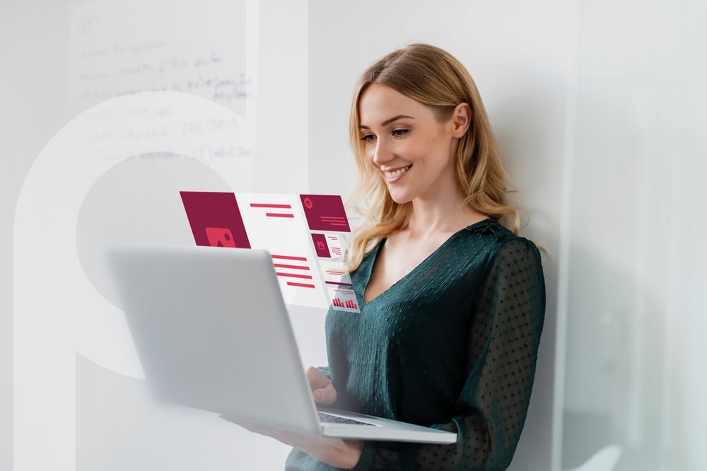Smiling blonde woman holding a laptop displaying digital elements. She is leaning against a white wall.